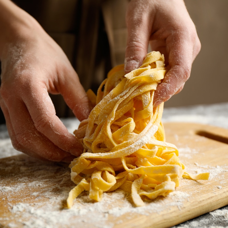 Pâtes fraîches maison : préparation de tagliatelles Mains enfarinées manipulant des tagliatelles fraîches et jaunes, déposées sur une planche à découper en bois couverte de farine.